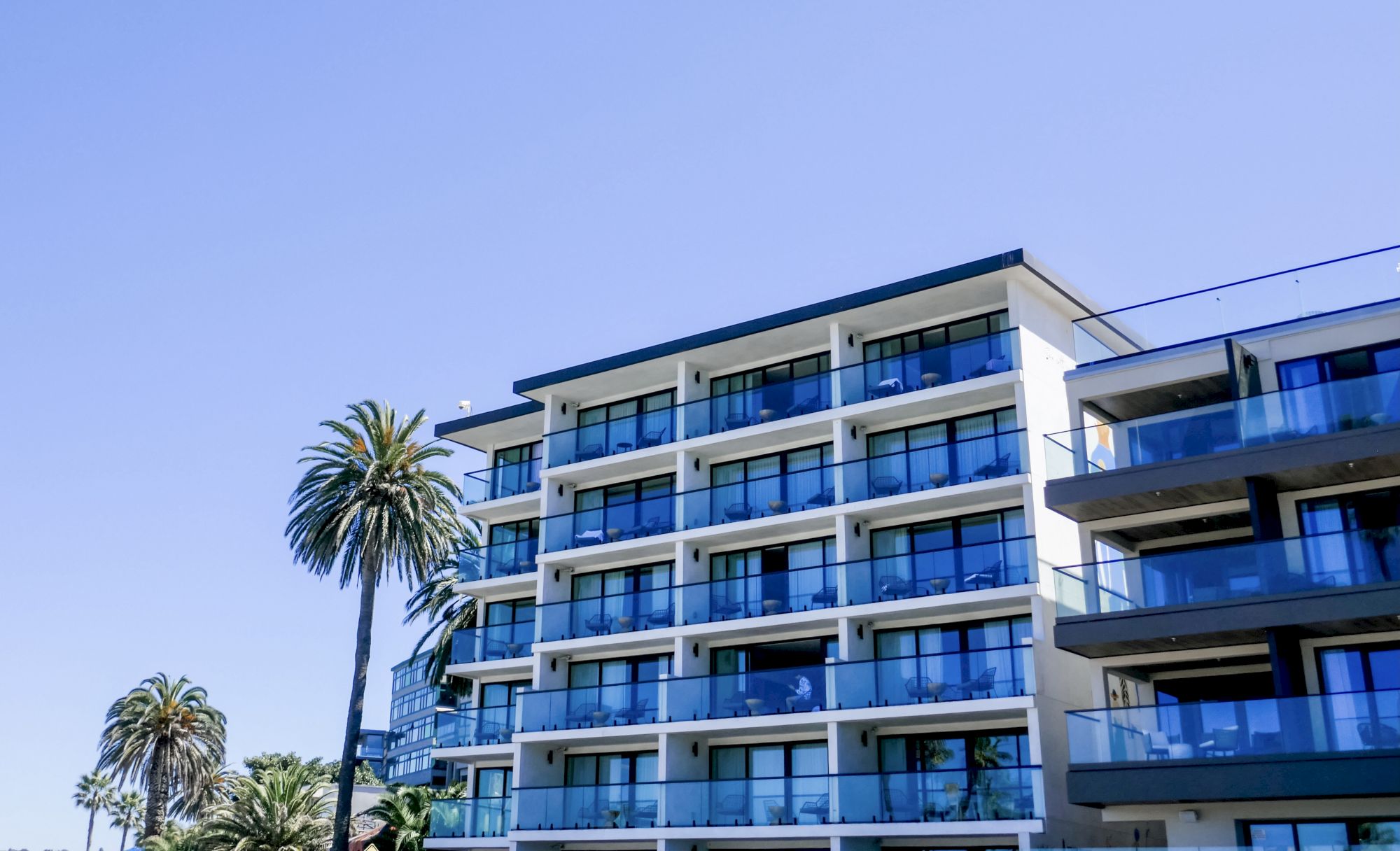 A modern, multi-story apartment building with glass balconies, palm trees, and a clear blue sky.