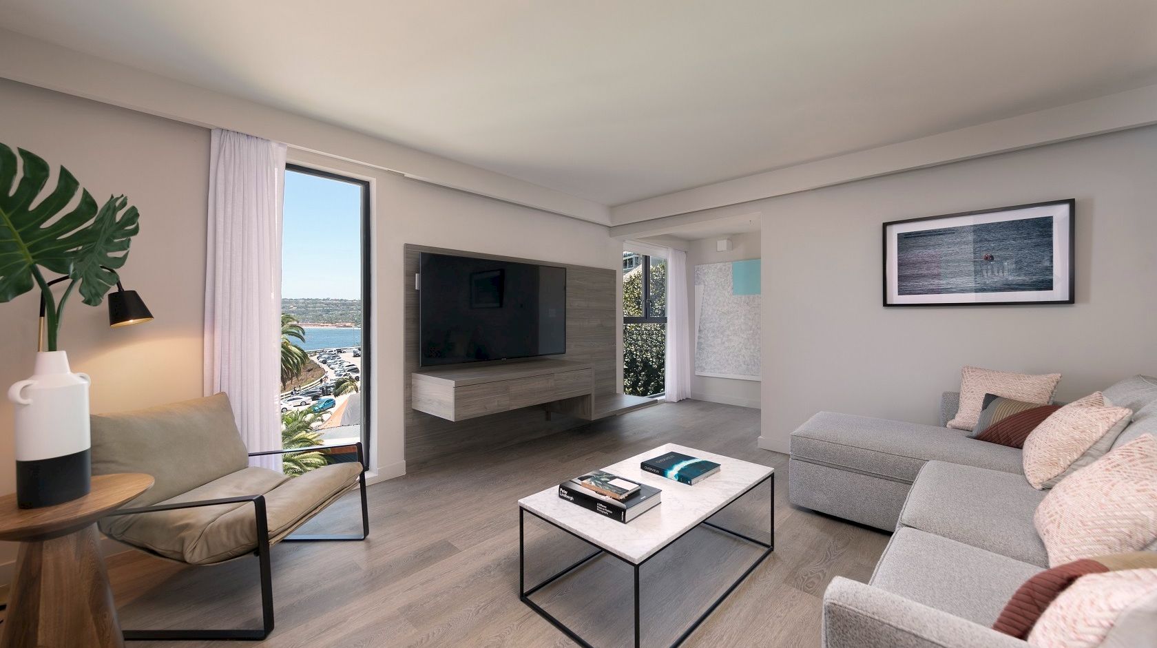 A modern coastal living room with a gray sectional, glass coffee table, wall-mounted TV, and a view of the sea through large windows, airy and bright.