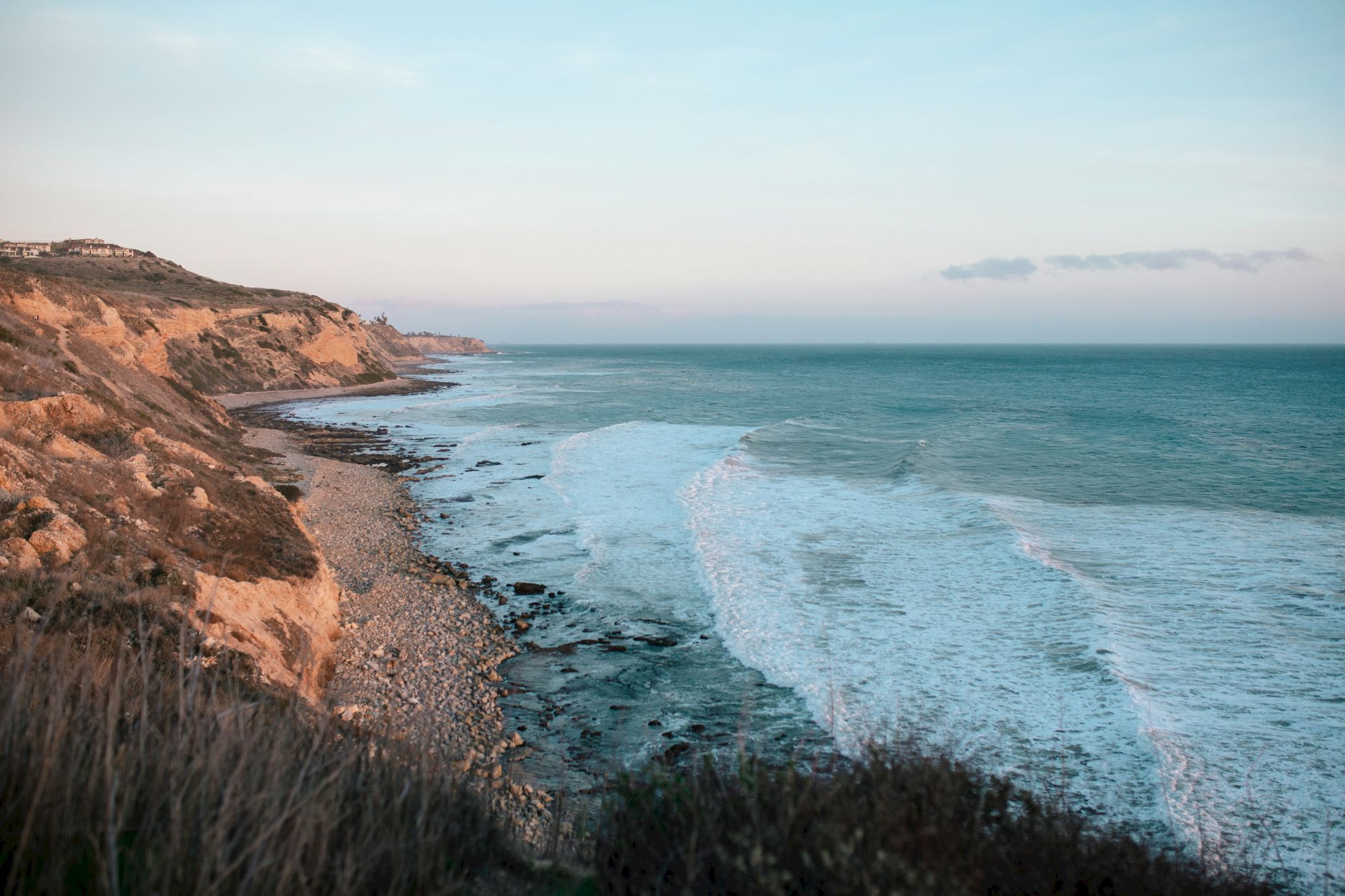 A coastal cliff overlooks a rocky shoreline with waves crashing onto the beach as the sun sets in a tranquil, expansive sea.