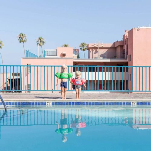 A sunny resort pool scene with lounge chairs, a bright pink building, palm trees, and guests relaxing by the water, vacation vibes abound.
