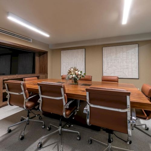 A modern home office with a long wooden desk, computer setup, shelving, and a window letting in natural light.