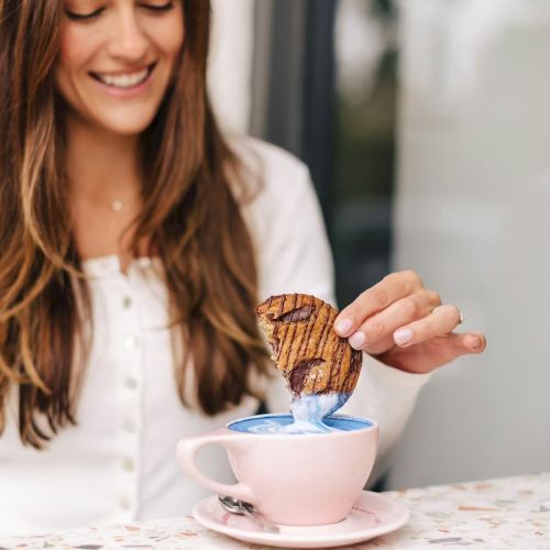 A young woman smiles while pouring tea into a pink cup perched on a saucer, with a pink plate of cookies nearby, bright and cozy.