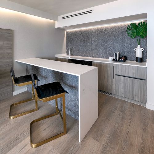 A modern kitchen with a white island, bar stools, wood accents, light wood flooring, and a potted plant on the counter.