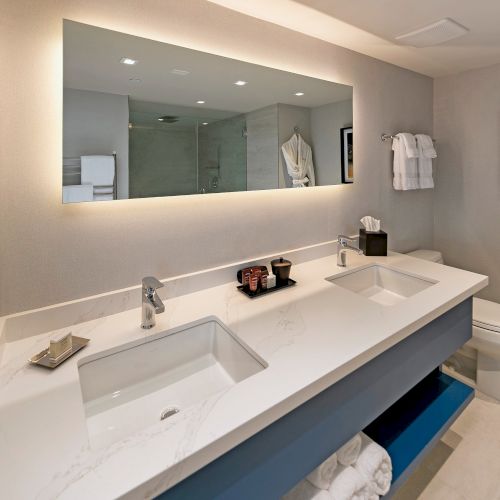 A modern bathroom with a long white counter, double sinks, blue accents, a large wall mirror, soft lighting, and neatly arranged towels.