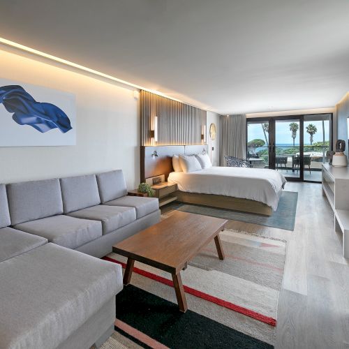 A modern hotel suite with a gray sectional, wooden coffee table, king bed, contemporary art, and a balcony view, bright and airy throughout.