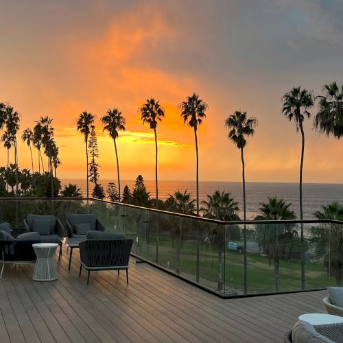 Sunset over a coastal deck with palm trees, glass railing, and outdoor seating silhouettes, calm ocean in the background.