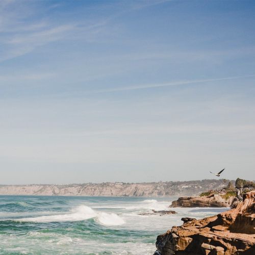 Waves crash on a rocky shoreline under a blue sky, with white foam and a tranquil sea meeting sunlit rocks at the shore.