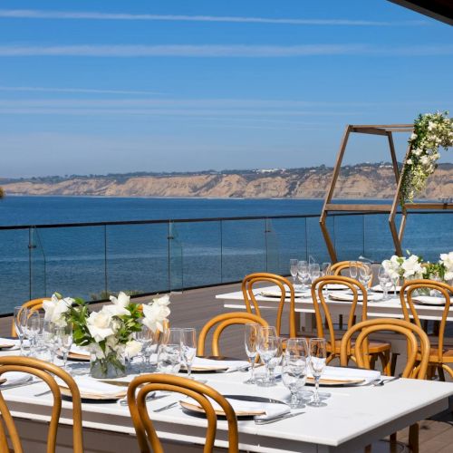 A seaside restaurant with white umbrellas, tables, and chairs along a rocky shore, calm blue water, and a clear sky. ending the sentence.