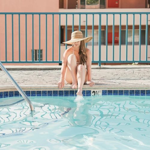 A person stands at the edge of a pool, wearing a swimsuit, with a fence and balcony in the background, a sunny outdoor scene.