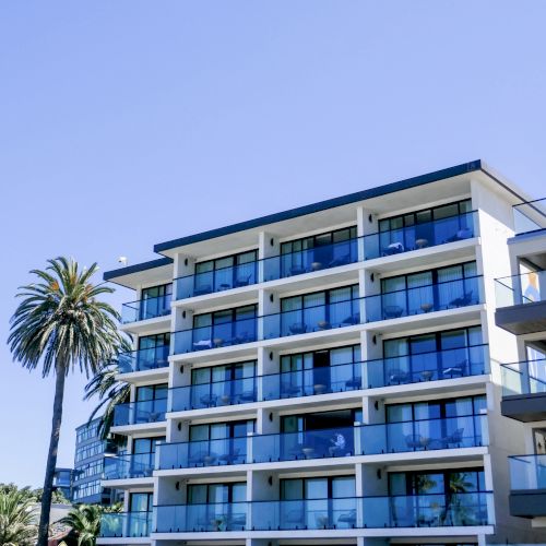 A blue multi-story building with glass balconies and palm trees in front, under a clear sky.