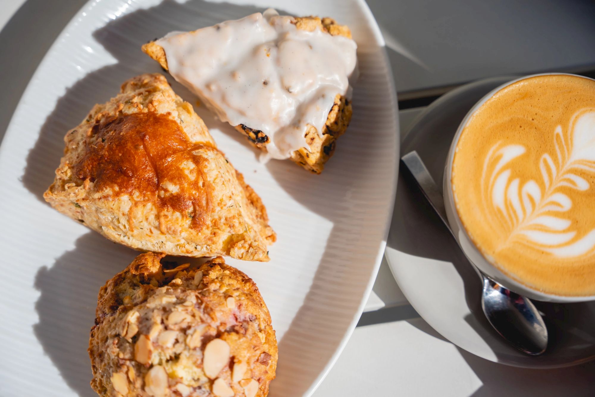 A plate with three pastries (almond/toasted nuts) and a cup of latte with latte art&mdash;sunlit, cozy cafe vibes.