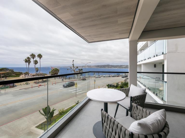 Balcony view over a street to the sea with a round table and cushioned chairs, palm trees, and a cloudy sky, modern glass railing.