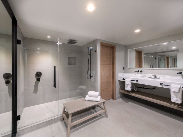 A sleek modern bathroom with a glass-walled shower, gray tiles, a wooden bench, and a long double sink vanity with large mirror and towels (ends with period).