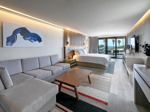 A modern hotel suite with a gray sectional sofa, wooden coffee table, and a king bed; stylish artwork, warm lighting, and a balcony view.