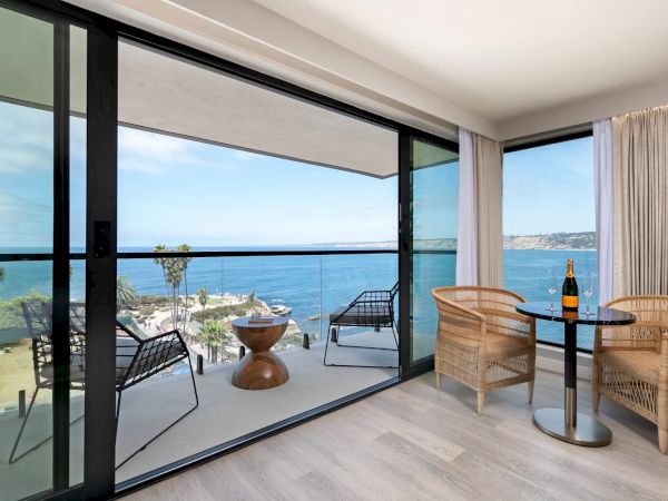 A modern hotel room with a glass sliding door opening to a balcony, overlooking the ocean, with wicker chairs, a small table, and a bottle on top, bright and airy atmosphere.