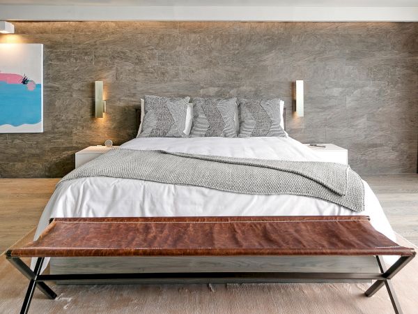 A modern bedroom with a large bed, gray bedding, a wooden bench at the foot, textured wall, and minimal decor.