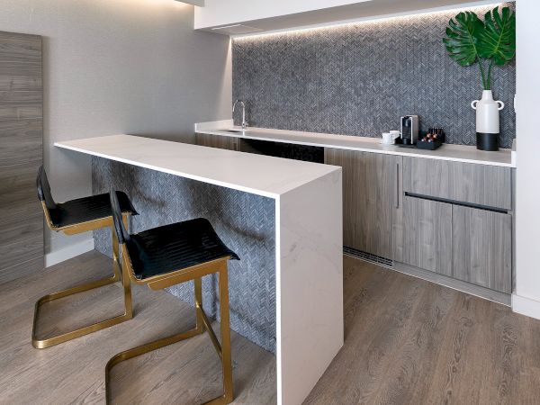A modern kitchenette with a light wood island, bar seating, grey cabinetry, a textured backsplash, and subtle under-cabinet lighting.