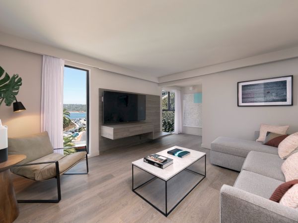 A bright, modern living room with a gray sectional, a sleek coffee table, a wall-mounted TV, and large windows revealing blue skies and greenery outside.