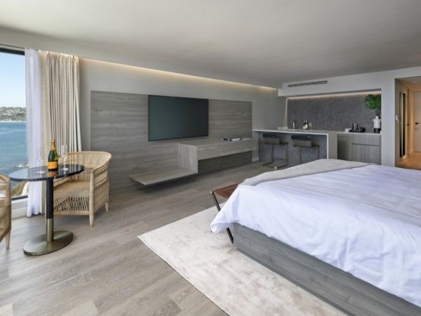 A sleek hotel room with a king bed, wall-mounted TV, a seating area by a window, a small kitchenette, and a light gray wooden theme.