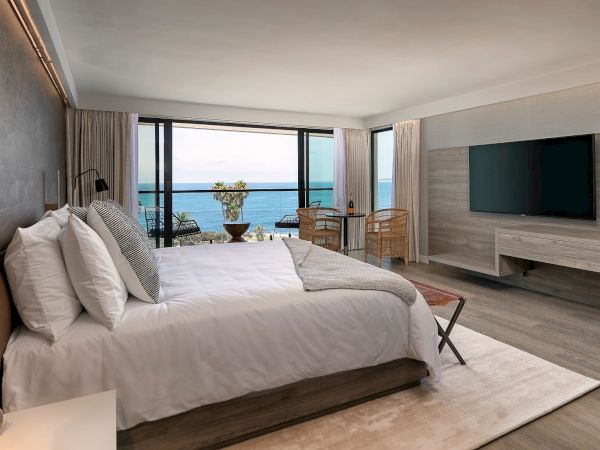 A modern hotel room with a large bed, light wood furniture, a wall-mounted TV, and a balcony view of the ocean. The scene feels airy and luxurious.