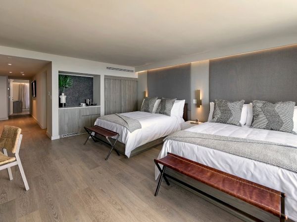 A modern hotel room with two large beds, gray upholstered headboards, soft lighting, a bench at each bed, a seating area, and a hallway leading to a kitchenette.