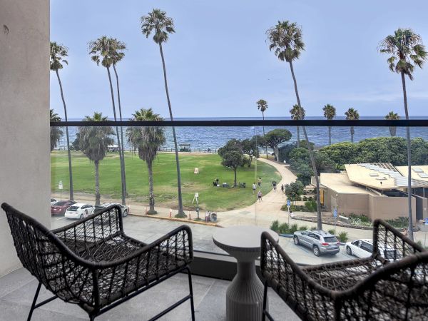 A sunny balcony view over a coastal park with palm trees, a path to the beach, and seating for two overlooking the ocean.