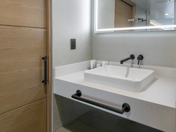 A modern bathroom with a wooden door, a rectangular vessel sink, a floating vanity with a black towel bar, and a backlit mirror.