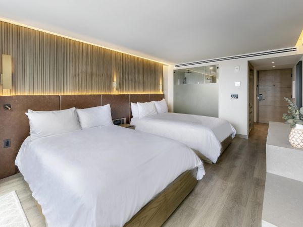A modern hotel room with two large white beds, soft lighting, a wooden headboard, light wood floors, and stylish decor including a vase on a pedestal.