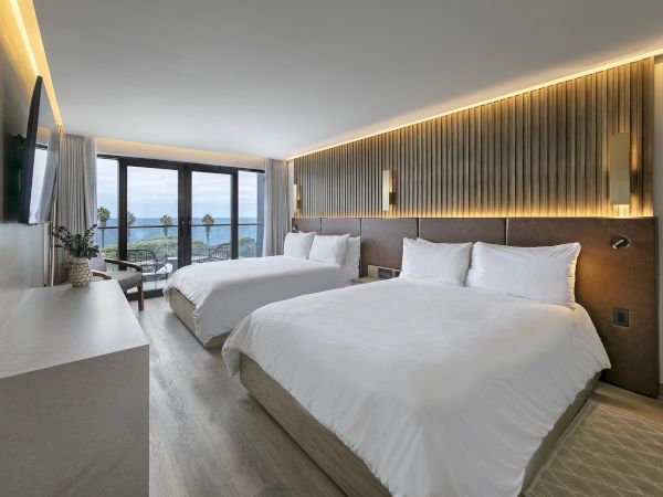 Two plush double beds in a bright, modern hotel room with a desk, wall TV, and a balcony view of the water&mdash;sleek and inviting.