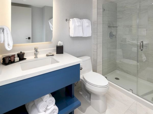 Small modern bathroom with a blue vanity, white countertop, double sink, wall mirror, hanging towels, and a glass-walled shower.