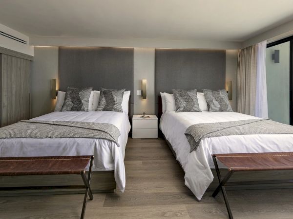 A bright hotel room with two queen beds, grey headboards, matching bedding, two benches at foot, nightstands, and a balcony ocean view.