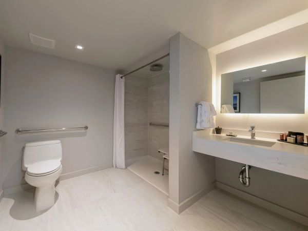 A clean, accessible hotel-style bathroom with a toilet, walk-in shower, and a wide vanity mirror with a countertop sink, all in light tones.