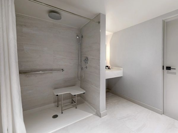 A modern, accessible bathroom with a tiled shower area, a foldable seating bench, a handheld shower, and a separate sink vanity across, all in light gray tones.