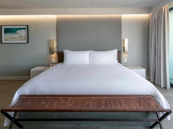 A modern hotel bedroom with a large white bed, two nightstands, wall sconces, a bench at the foot, soft lighting, and a framed seaside picture on the left.