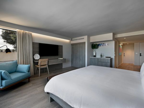 A modern hotel room with a large bed, a blue sofa, a wall-mounted TV, a small desk, and a kitchenette area, plus natural light from a window.