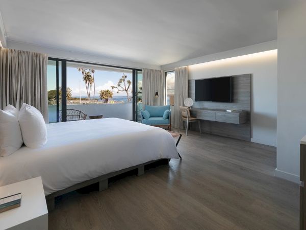 A modern hotel room with a large bed, light wood floors, and a seating area by a wall-mounted TV, overlooking a beachfront view with palm trees.