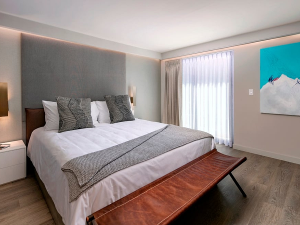 A modern hotel bedroom with a large bed, gray upholstered headboard, bedside lamps, a bench at the foot, and a colorful abstract painting by the window.
