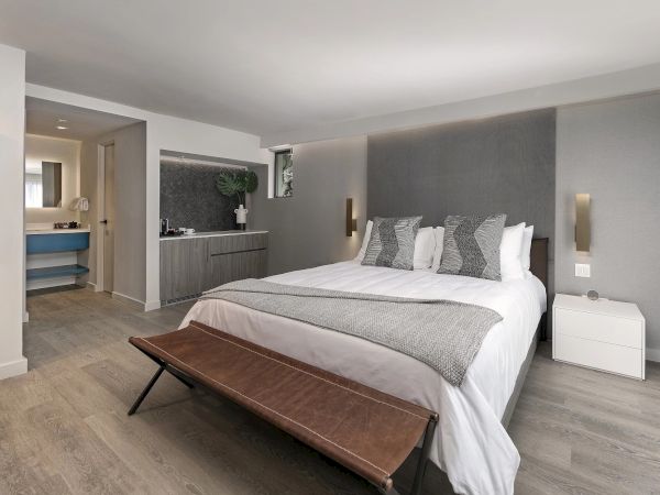 A modern hotel-style bedroom with a king bed, gray upholstered headboard, white bedding, two nightstands, a bench at foot, and a small kitchenette area in the back corner.