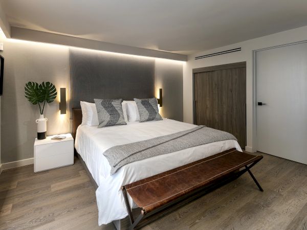 A modern hotel bedroom with a large bed, grey and white tones, two nightstands, a bench at the foot, and ambient wall lighting.