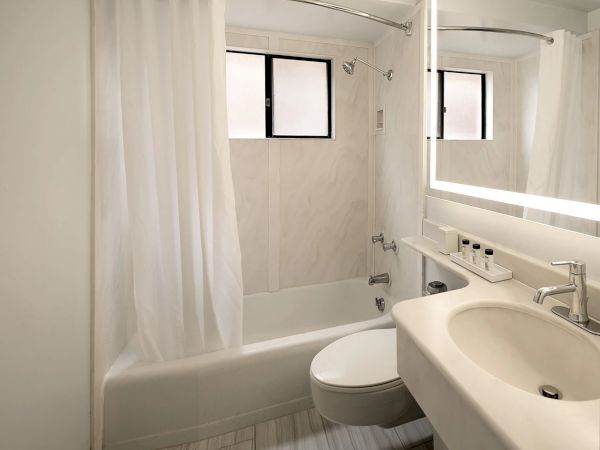 A compact modern bathroom with a bathtub/shower combo, white tile, frosted curtains, a wall mirror with built-in lighting, and a sink with fixtures.