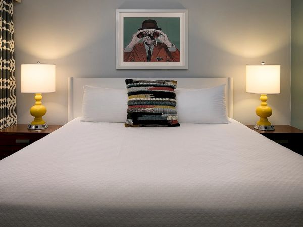 A neatly made bed with two yellow bedside lamps, a stack of colorful pillows, and a framed photo above the headboard, in a calm bedroom.