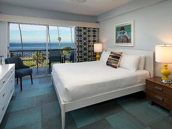A bright hotel room with a large bed, nightstands, lamps, a dresser, seating area, and a balcony view of the ocean through sliding glass doors.
