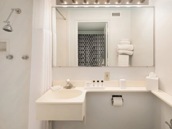 A white bathroom with a wall mirror, floating sink, toilet paper holder, stacked towels, and a bright, rowed vanity light above the mirror.