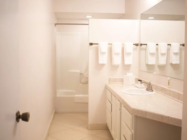 A clean, white bathroom with a single sink vanity, lots of towels on a rack, and a shower/tub at the far end, bright and minimalist.