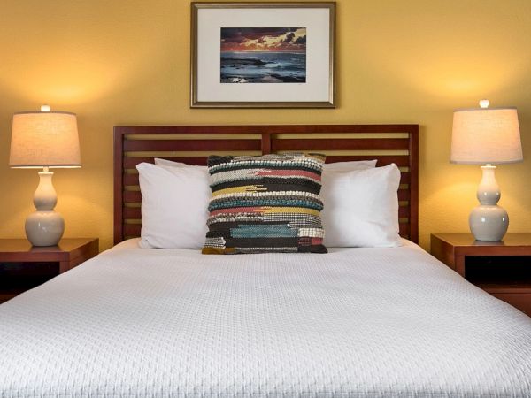 A neatly made bed with a white quilt, two nightstands, matching lamps, a colorful decorative pillow, and a framed seascape above the headboard.