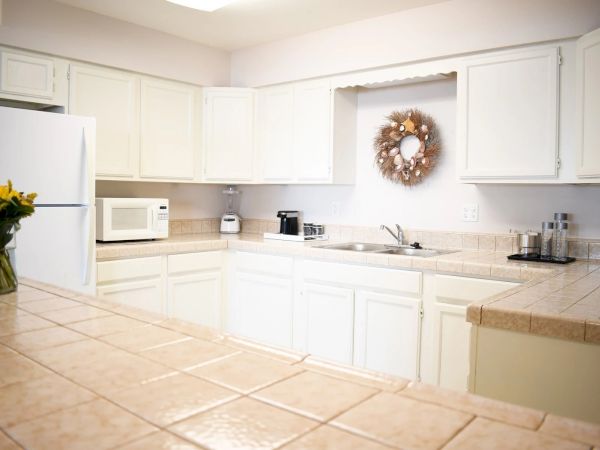 Spacious white kitchen with tiled counters, white cabinets, a refrigerator, microwave, coffee maker, vase of flowers, and a decorative wreath on the wall.