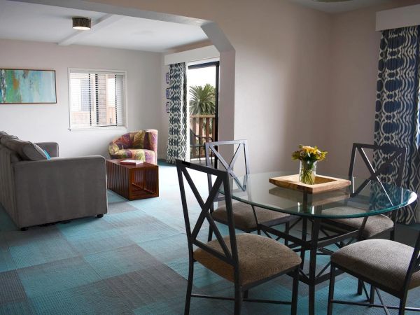 A cozy living and dining area in a bright apartment: teal carpet, a gray sofa, wooden chair, glass dining table with metal chairs, and a small vase of yellow flowers.