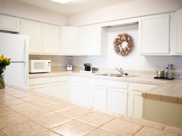 A bright, U-shaped kitchen with white cabinets, tiled countertops, a toaster, microwave, coffee maker, sink, and a decorative wreath on the wall.