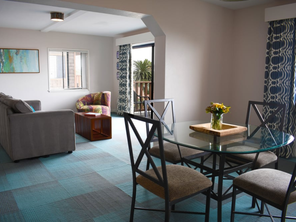 A bright living and dining area with blue patterned carpet, a glass round table, four chairs, a gray sofa, armchairs, and a balcony view.
