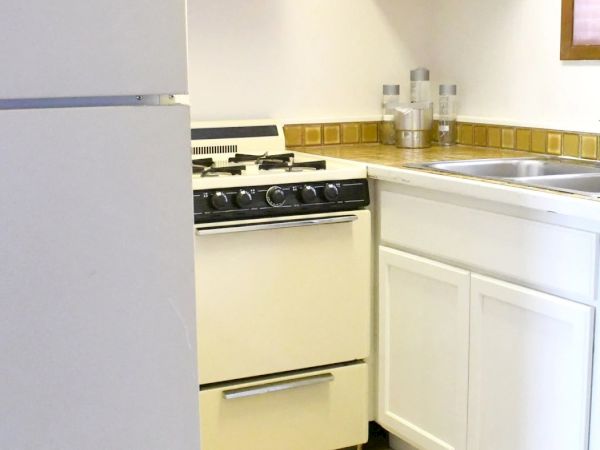 A small, dated kitchen with a white fridge, stove, and cabinets, plus a tiled backsplash and sink counter.